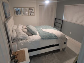 A bedroom with a bed, pillows, a blanket, a bedside table with a lamp, a wall-mounted artwork, a closet, and a window with blinds. - Cozy Coastal Suite  (Bradenton)