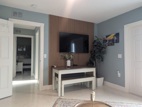 A room with a television mounted on a wall, a wooden console table, a potted plant, and a door leading to another room. - Cozy Coastal Suite  (Bradenton)