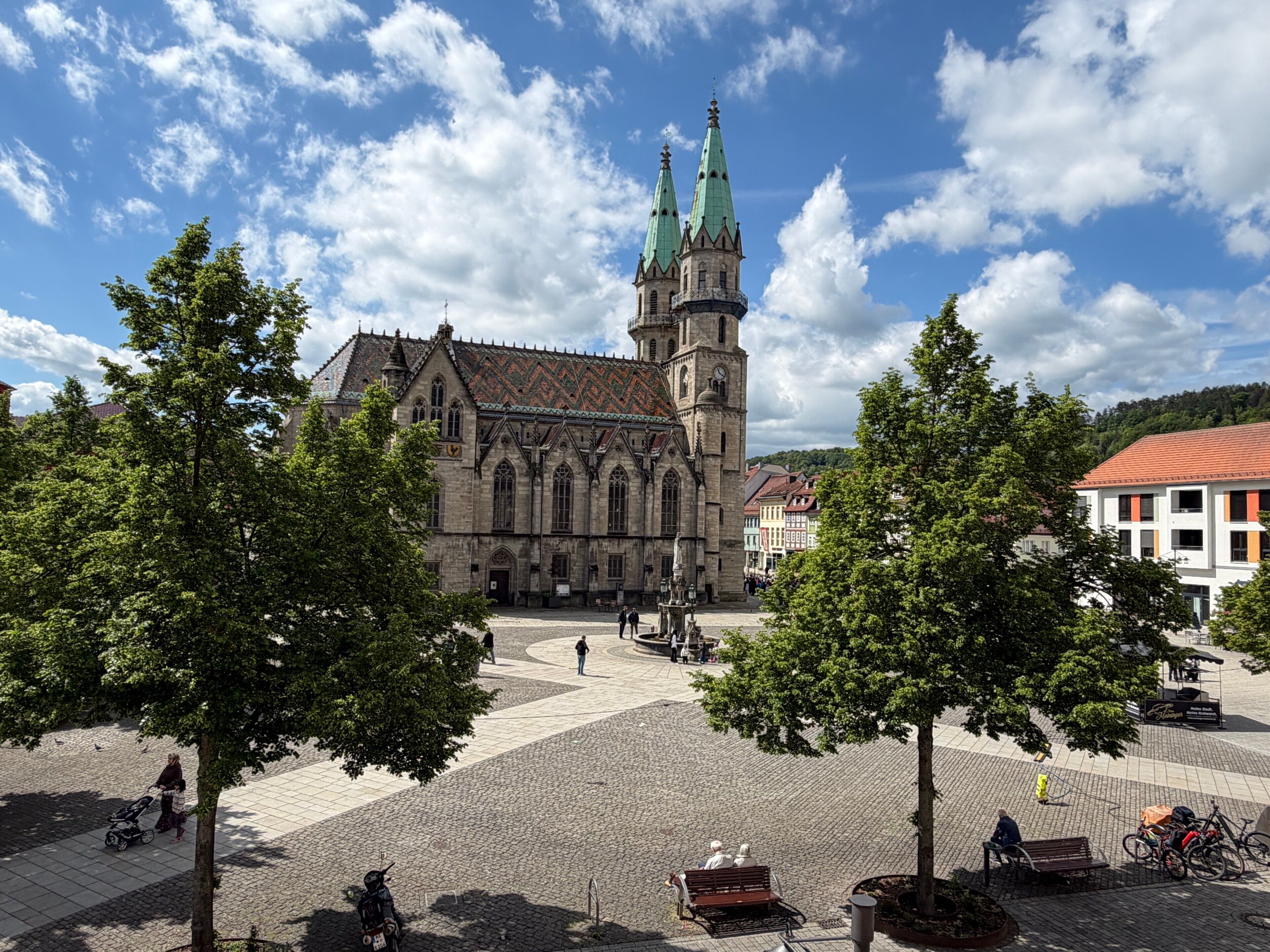 Eine historische Kirche mit einem markanten Turm und einem gepflasterten Platz mit Bänken und Bäumen.