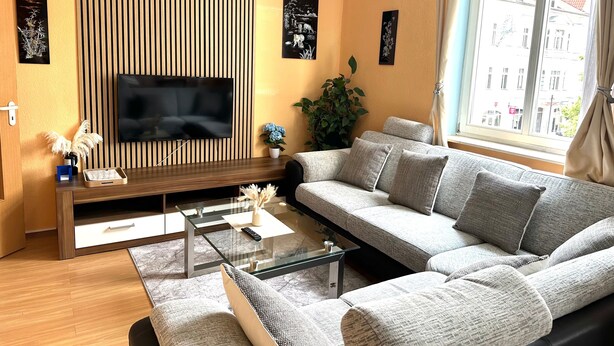 Una sala de estar con un sofá modular, una mesa de centro de vidrio, un televisor de pantalla plana montado en una pared de paneles de madera y una planta en maceta.