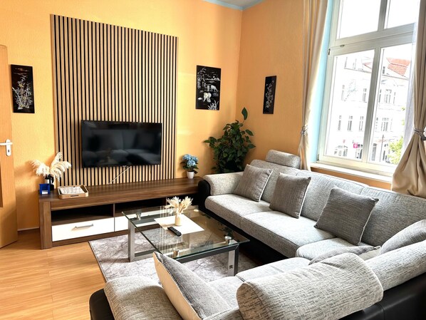 Een woonkamer met een hoekbank, een glazen salontafel, een flatscreen televisie bevestigd aan een houten wandpaneel, en een potplant.
