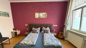 Un dormitorio con una cama doble, una mesita de noche con lámpara, una guitarra y una ventana con cortinas.
