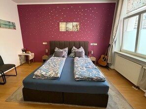 Een slaapkamer met een tweepersoonsbed, een klein tafeltje met een lamp, een gitaar en een raam met gordijnen.