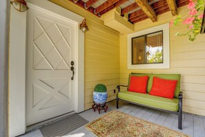 A room with a white door, a green sofa with red cushions, a blue vase, and a window with blinds.