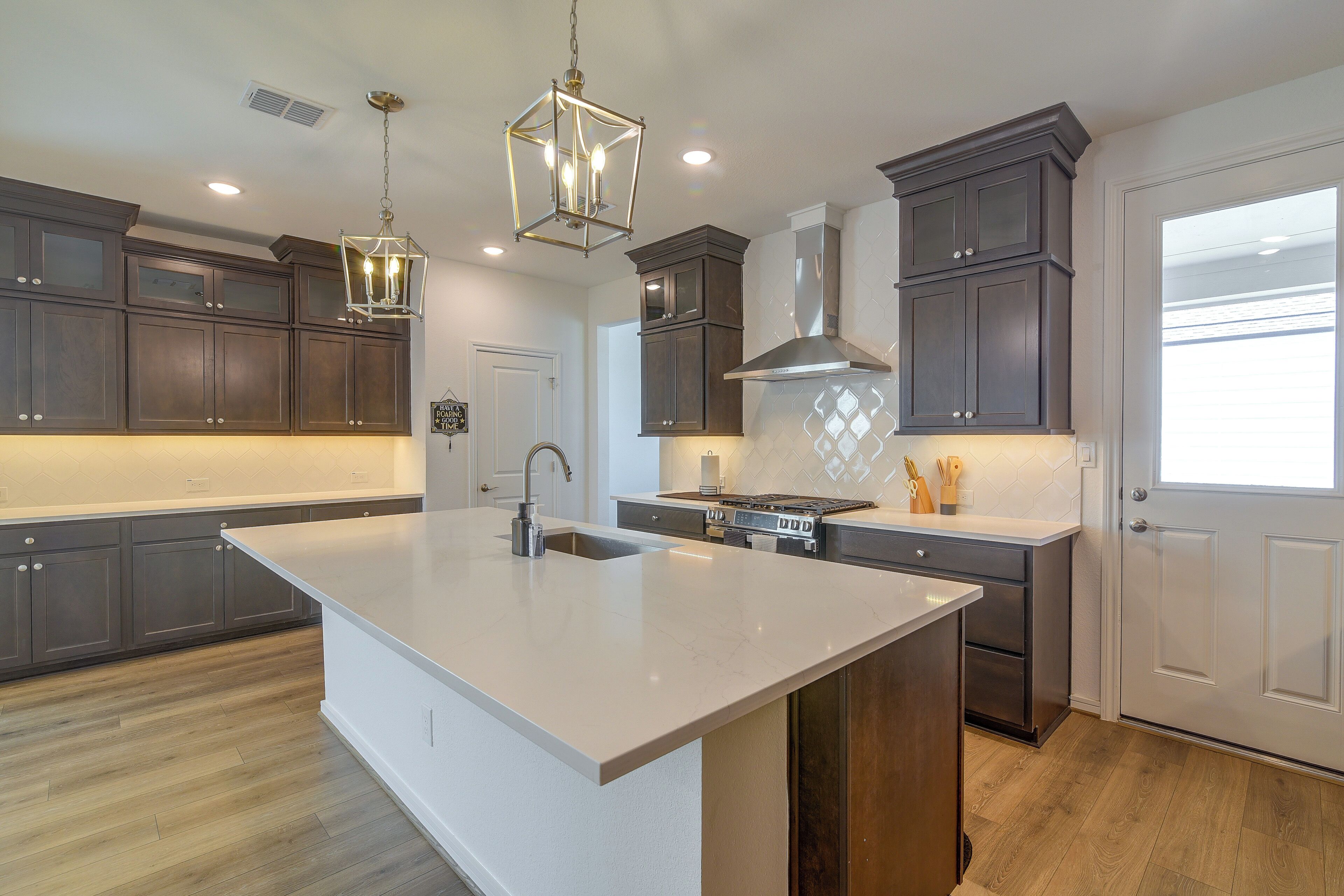 A modern kitchen with dark wood cabinets, a large island with a sink, stainless steel appliances, and pendant lighting.
