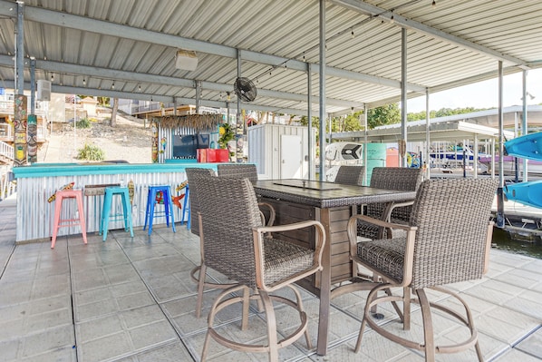 Een terras met een tafel en stoelen, een ventilator en zicht op boten op de achtergrond.