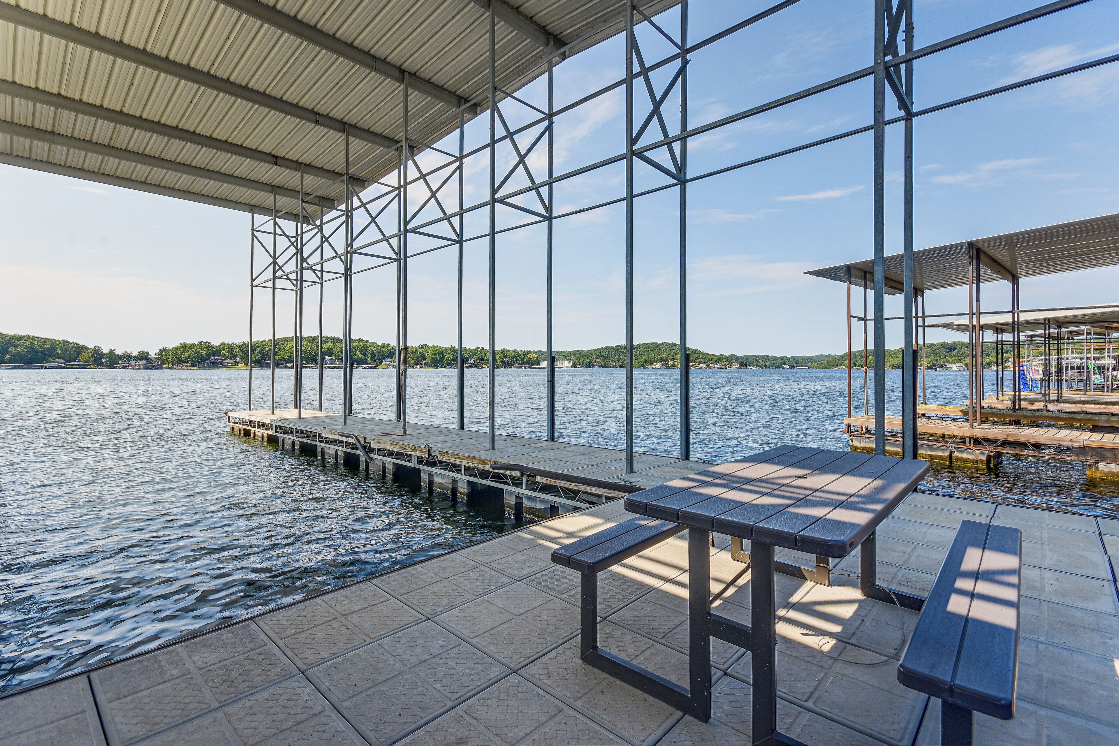 A covered dock area with a table and benches, overlooking a calm body of water with a distant shoreline.