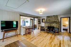 Un soggiorno con televisione su un mobile, un camino in pietra, pavimento in legno e una zona cucina visibile tramite un'apertura.