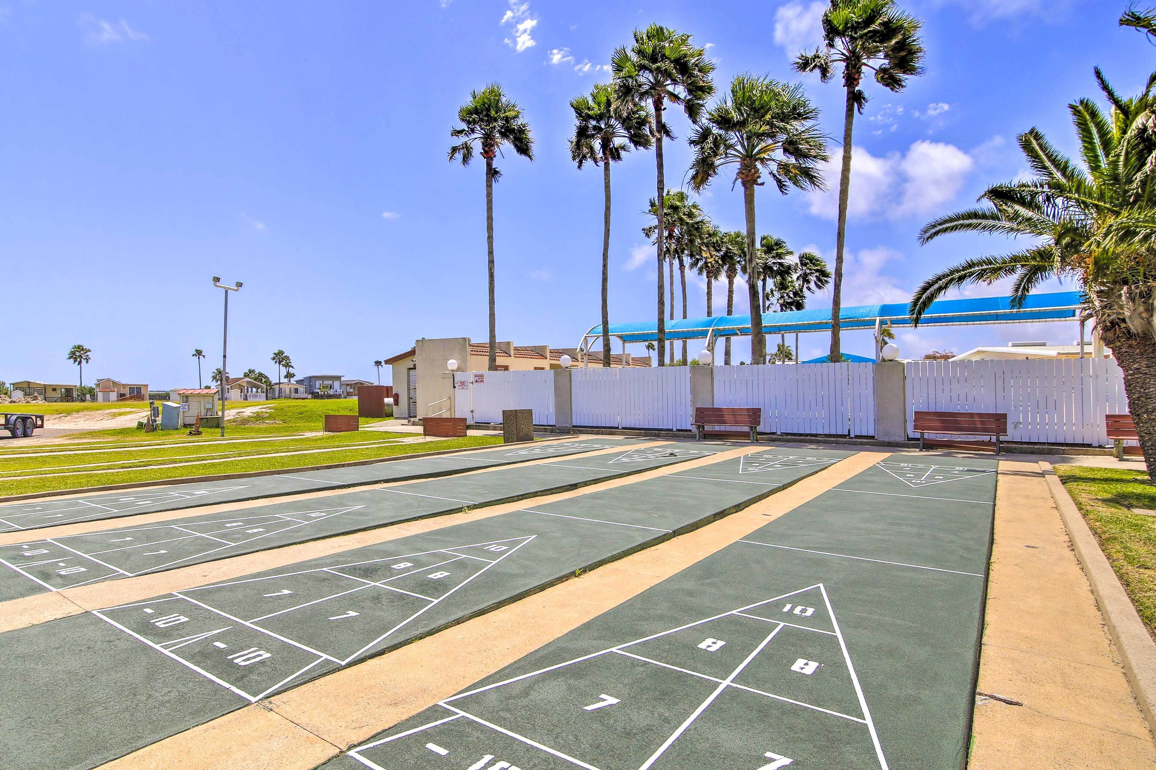 Ein Shuffleboard-Spielfeld mit nummerierten Zählfeldern, umgeben von Palmen und einem blauen Gebäude im Hintergrund.