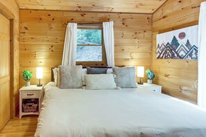 Un dormitorio de cabaña de madera con una cama, dos mesitas de noche, un ventanal con cortinas y un cuadro en la pared.