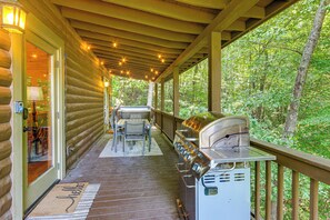 Terraza exterior techada con una parrilla, una mesa con sillas y barandilla de madera con vista a un bosque.