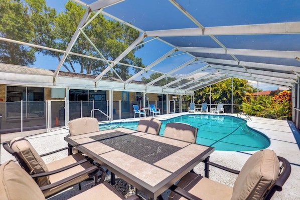 A covered outdoor pool area with a table and chairs, surrounded by glass walls and a view of trees and a building.