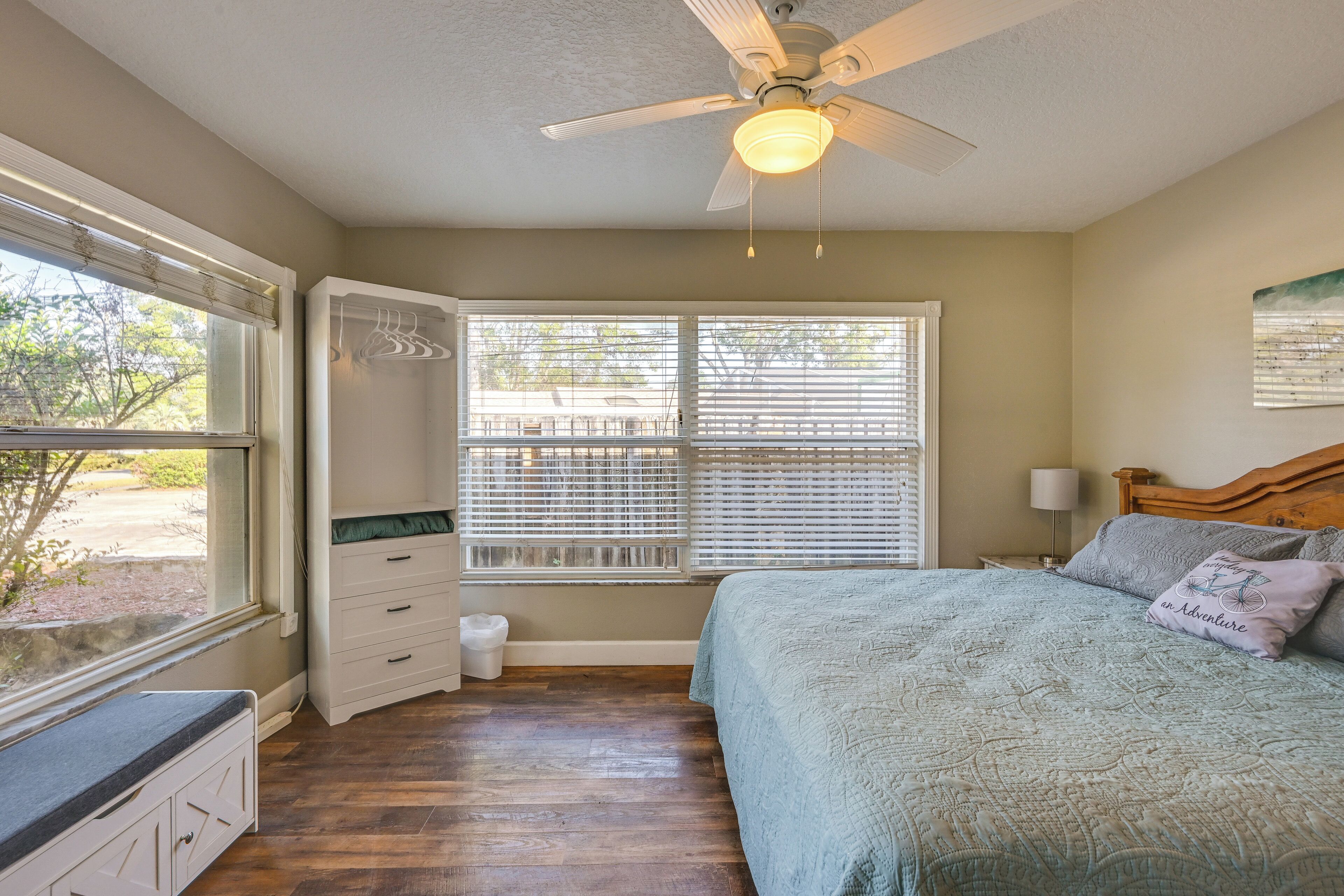 A bedroom with a bed, a ceiling fan, a window with blinds, a nightstand, and a bench by the window.