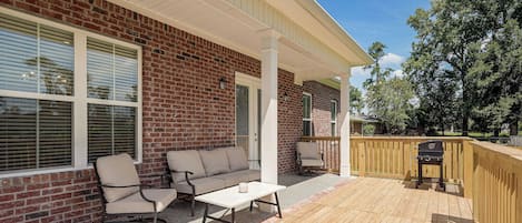 A covered patio with a brick wall, a wooden deck, outdoor furniture, and a grill.