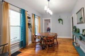 Un comedor con mesa y sillas de madera, una araña y vistas al exterior a través de una ventana con cortinas.