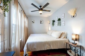 Un dormitorio con cama, ventilador de techo, mesita de noche con lámpara y obras de arte colgadas en la pared.