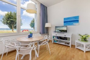 Una habitación con una mesa de comedor de madera, sillas blancas, un televisor sobre un mueble blanco y una ventana con vista a la playa.