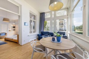 A modern room with a round dining table, white chairs, a blue sofa, and a large window with a view of buildings. - Beach bliss at Villa Agnes (Binz)