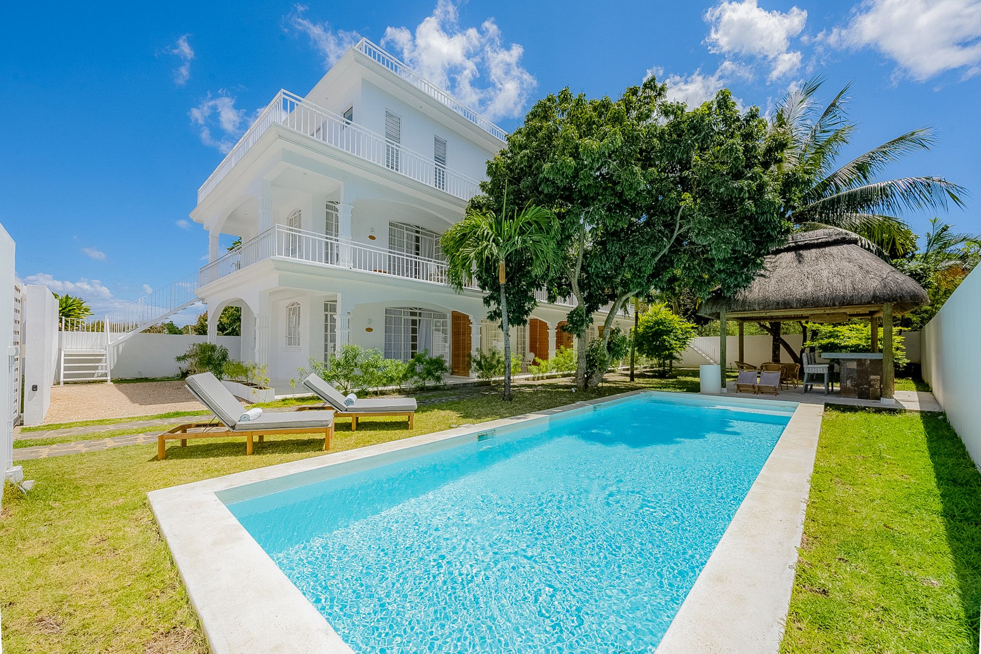 A two-story white building with a swimming pool, lounge chairs, and a thatched-roof structure.