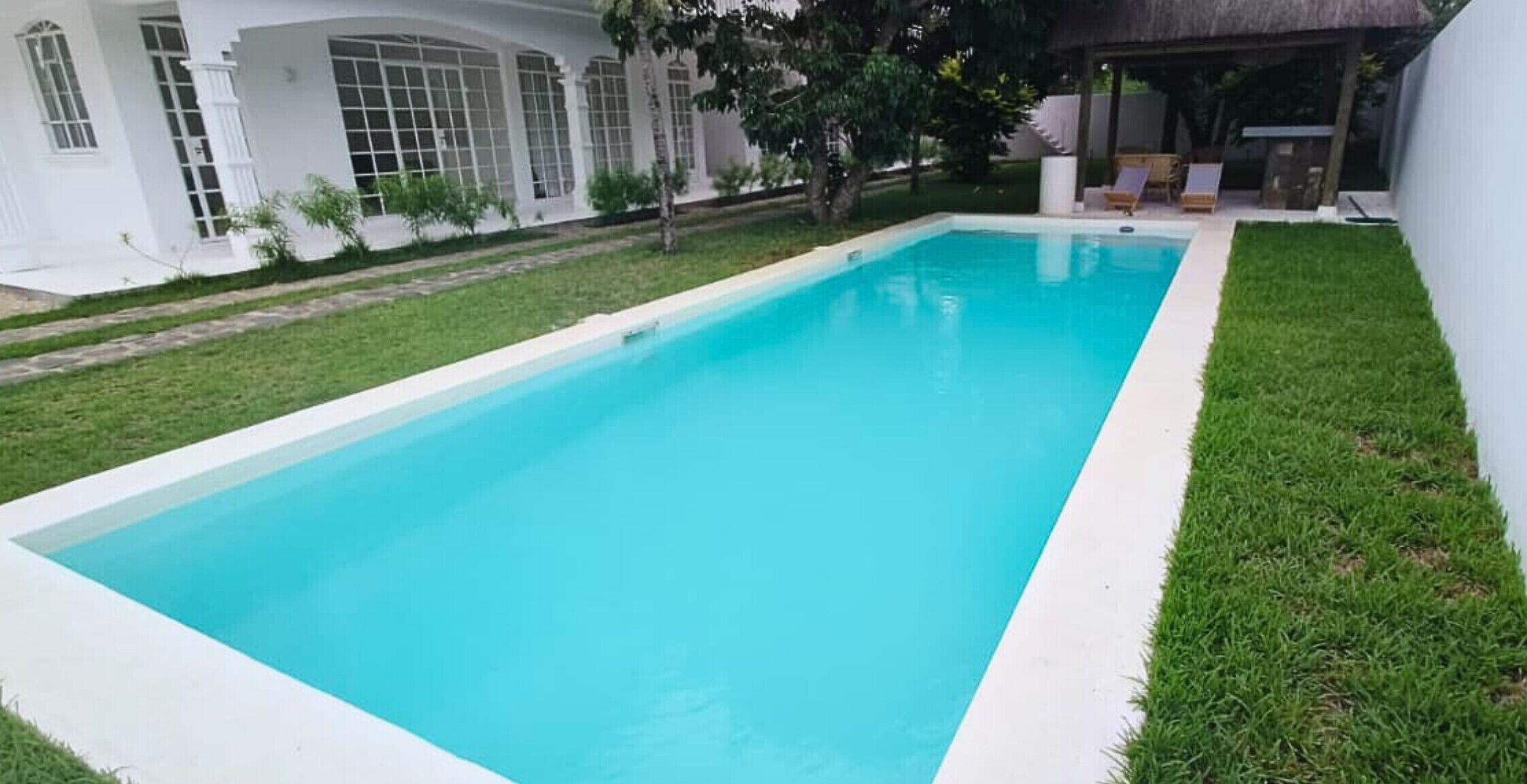 Ein rechteckiger Swimmingpool mit weißem Rand, umgeben von einer gepflegten Rasenfläche und einem Gebäude mit großen Fenstern, das in Weiß gehalten ist.