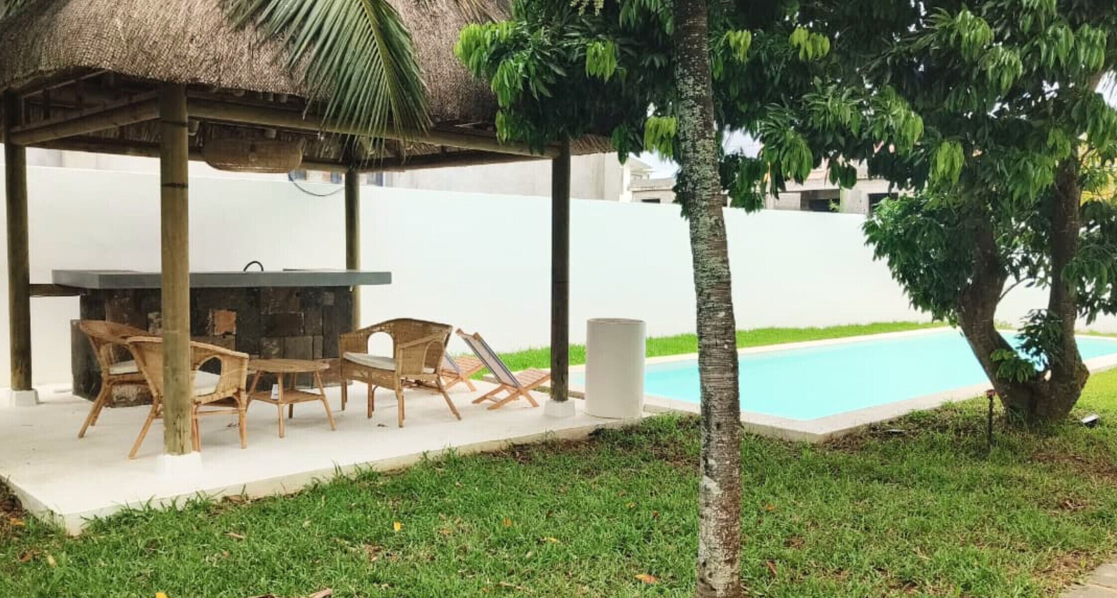 Un espace extérieur aménagé avec des sièges et une table, protégé par un auvent, ainsi qu’une piscine et un palmier.