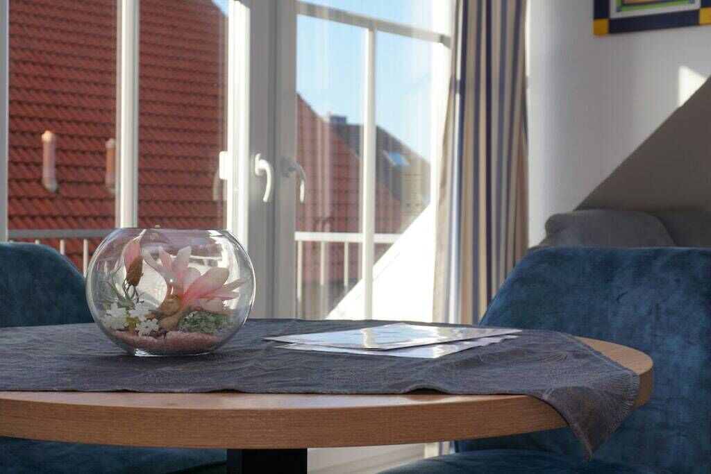 A round wooden table with a glass terrarium and papers on it, set against a blue couch and a window with a view of buildings.