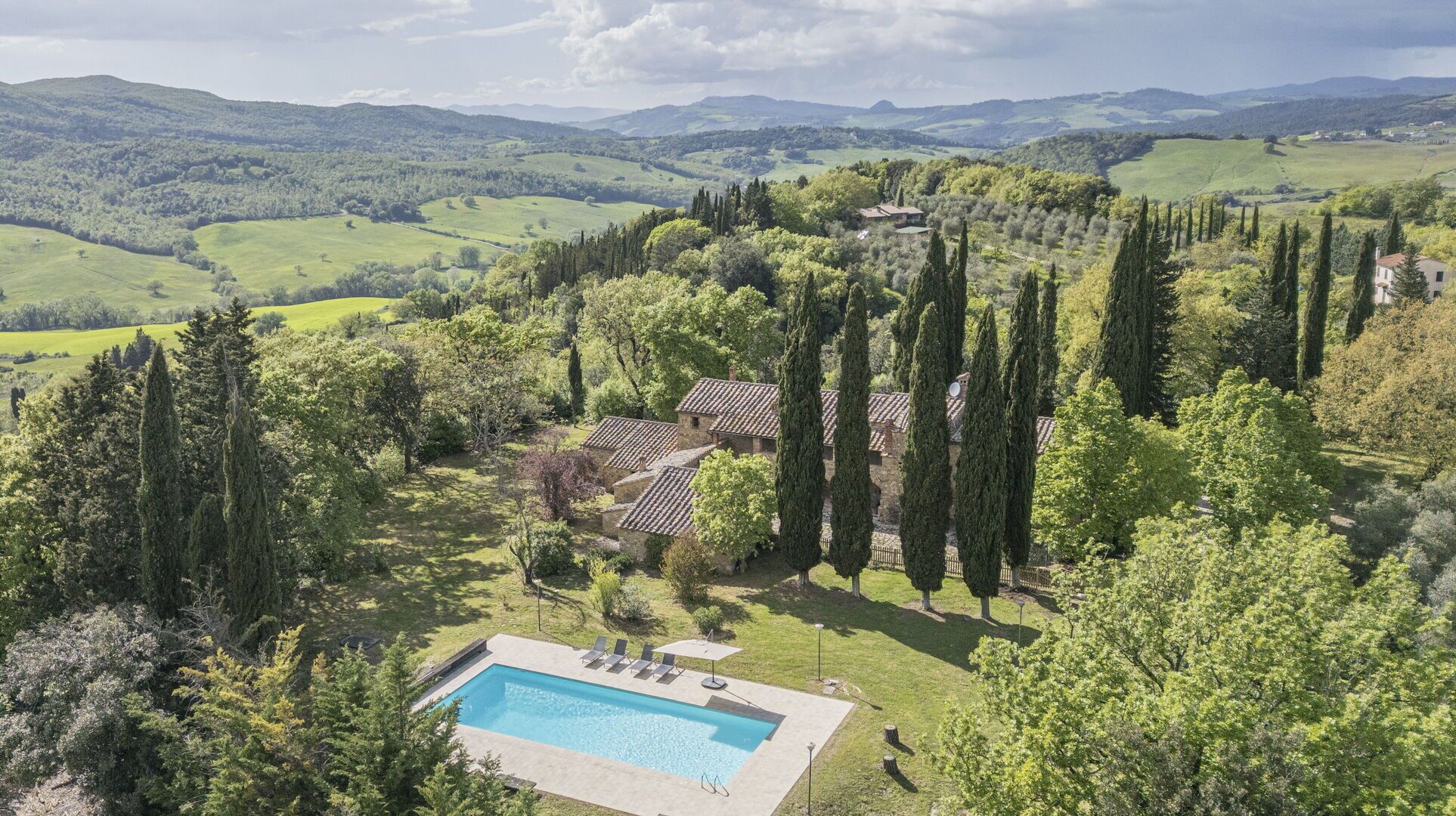 Ein Pool, umgeben von einem Garten mit hohen Bäumen, ein Steinhaus und Blick auf sanfte Hügel.