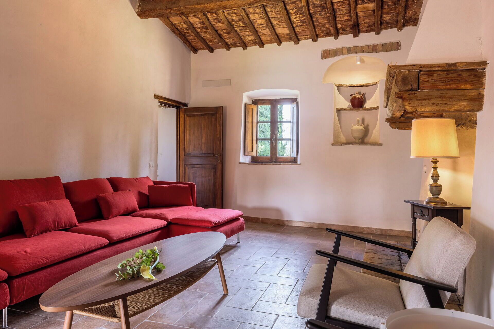 Una habitación con sofá rojo, una mesa de centro de madera, una silla, una lámpara y un ventanal con vistas al verdor.