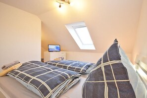 Une chambre avec un plafond en pente, un puits de lumière, un lit recouvert d’une couverture à carreaux, une table de chevet avec une lampe et un téléviseur.