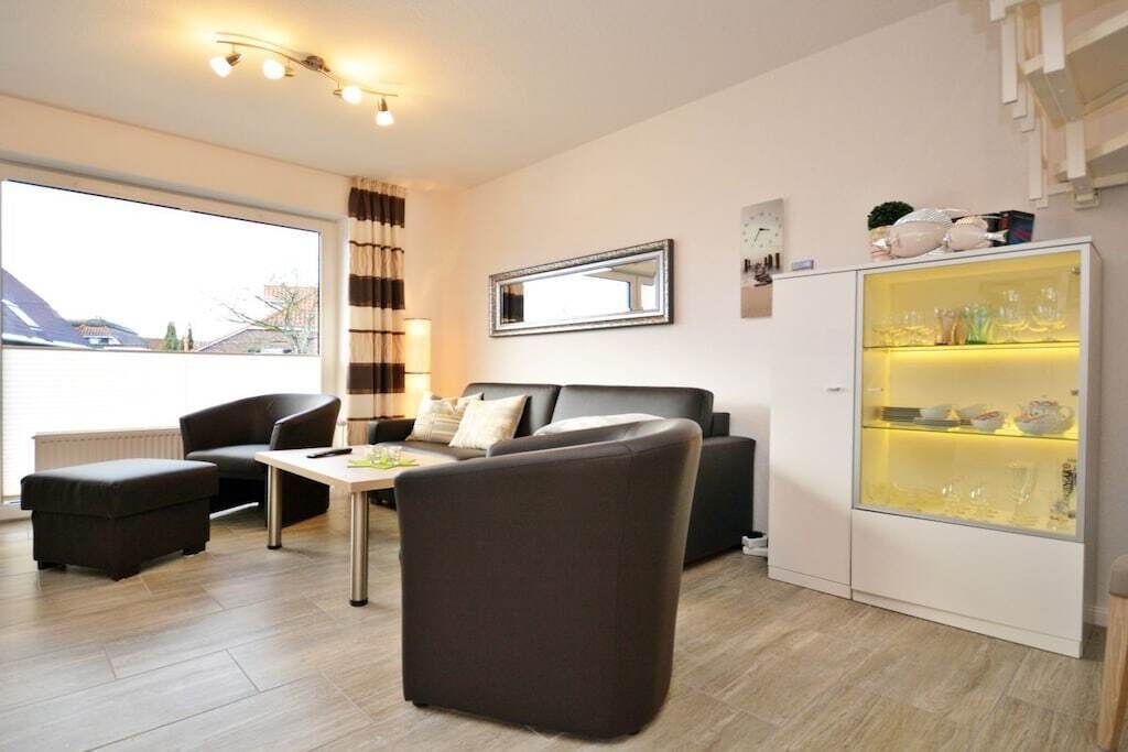Una sala moderna con sofá, sillón y reposapiés, un aparador de cristal con adornos, y un ventanal con cortinas.