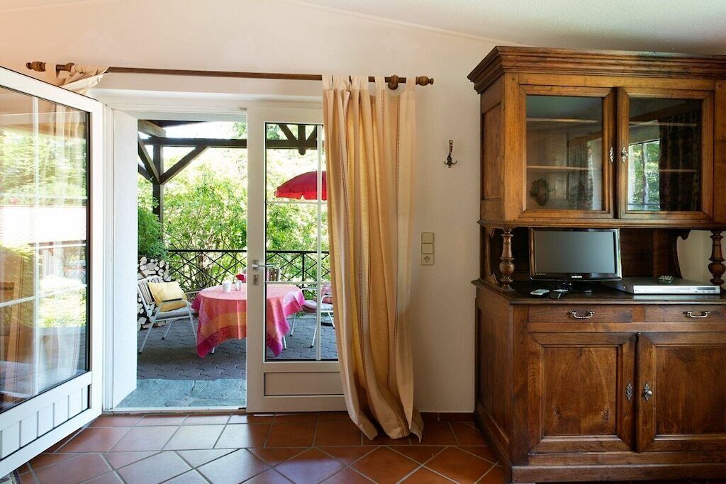Ein Zimmer mit einer Schiebeglastür, die auf eine Terrasse mit Tisch und Stühlen führt, einem Holzschrank mit Fernseher und einem teilweise gezogenen Vorhang.