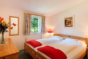 A double bed with red pillows, a bedside table with a vase of flowers, a window with curtains, and a poster on the wall. - Holiday apartment with 1 bedroom (Krumpendorf)