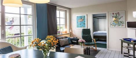 A room with a dining table, a sofa, a chair, and a bunk bed. There are windows, artwork on the walls, and a checkered floor.
