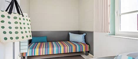 A small, cozy room with a striped bed, a window with striped curtains, and a polka-dotted bag hanging on the wall.