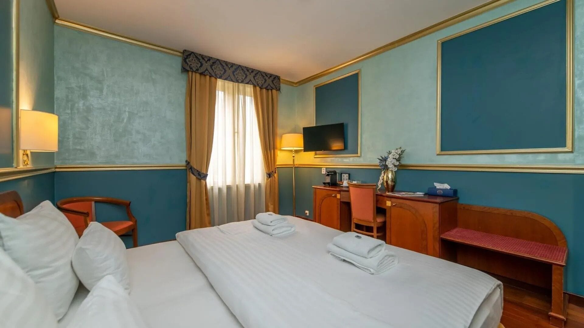 Ein Hotelzimmer mit einem großen Bett, einem Schreibtisch, einem Sessel, einem Fernseher und einem Fenster mit Vorhängen.