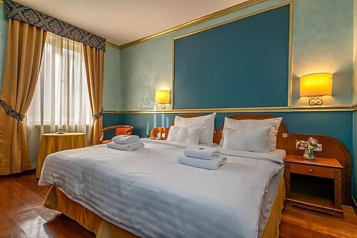 A hotel room with a large bed, two bedside tables, a nightstand, and a window with curtains.