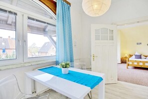 Ein Zimmer mit einem weißen Tisch, einem blauen Vorhang, einem Blumentopf, einem Bett mit Holz-Kopfteil und einer Tür mit Fenster.