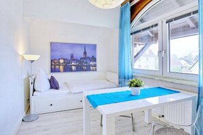 A modern room with a white sofa, a table with a blue tablecloth, a cityscape painting on the wall, and a view through a window. - Holiday apartment with 1 bedroom (Travemünde)