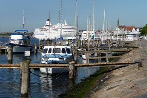 Ein Yachthafen mit vertäuten Booten, im Hintergrund ein großes Kreuzfahrtschiff und ein Holzsteg, der zu einer Stadt mit Gebäuden führt.
