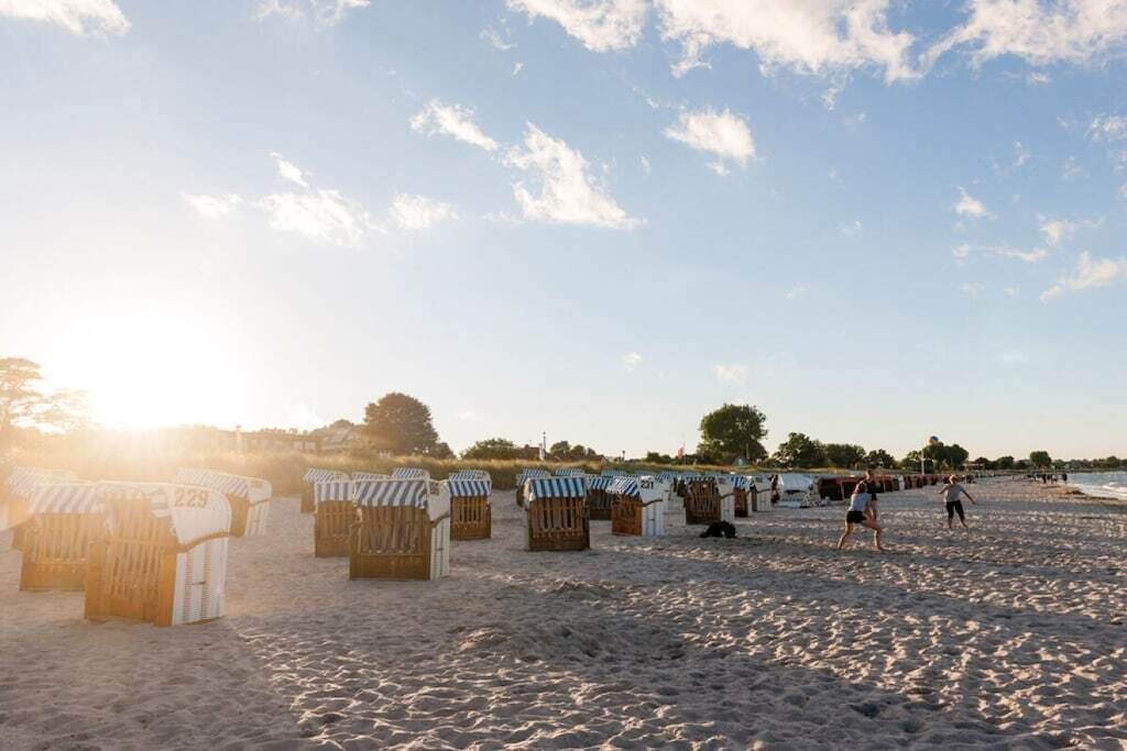 Ein Strand mit Reihen aus Holzstrandhäusern, von denen einige mit Nummern versehen sind, und ein paar Personen, die am Sand entlanggehen.