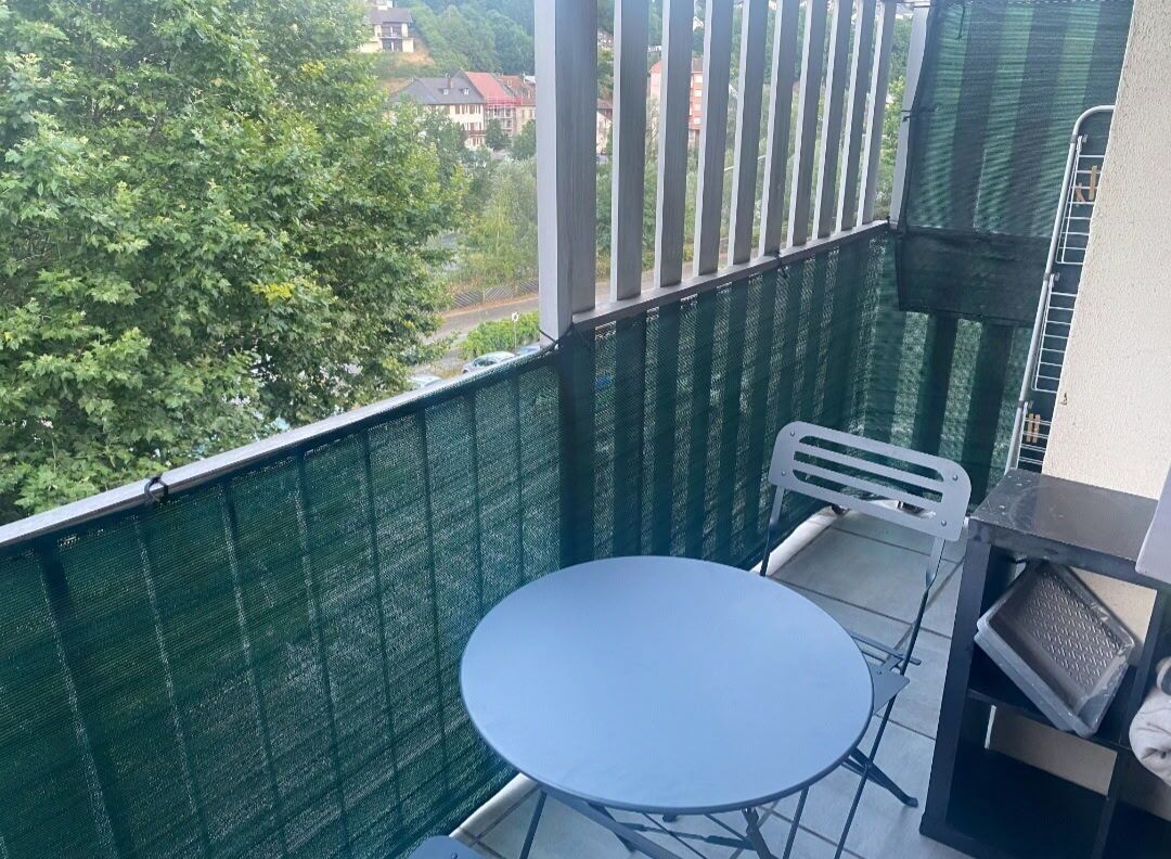 A balcony with a round table and a chair, a green mesh screen, and a view of trees and buildings.