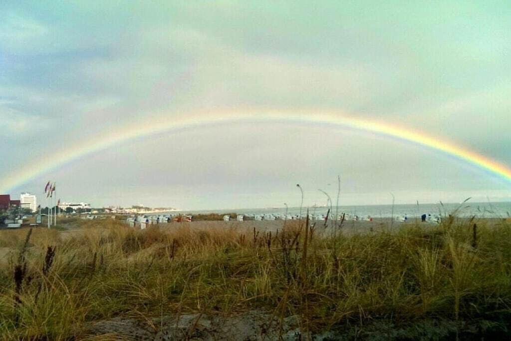 A beach with a rainbow, grassy dunes, and a distant view of buildings.