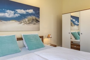 A hotel room with a bed, two pillows, a nightstand with a lamp, and a large painting of a beach scene on the wall. - Holiday apartment with 1 bedroom (Travemünde)