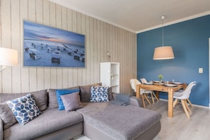 A modern living room with a grey sofa, a dining table with chairs, a painting of a beach scene, and a blue accent wall. - Holiday apartment with 1 bedroom (Travemünde)