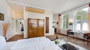 A hotel room with a large bed, a wooden wardrobe, a dining area with a table and chairs, and a view of the outside. - Villa Agnes Apartment 11 (Binz)