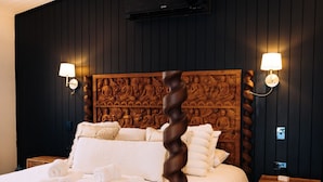 Uma cama de dossel com lençóis brancos e um cobertor amarelo, ladeada por duas mesas de cabeceira com luminárias fixadas na parede, em um quarto com painéis de madeira escura e um ventilador de teto.