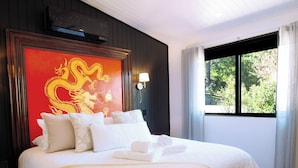 Quarto de hotel com uma cama grande, um ventilador de teto, uma obra de arte de dragão vermelho e uma janela com vista para vegetação.