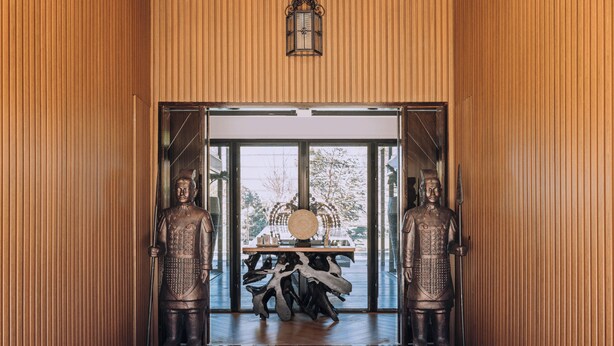 Interior em madeira com duas estátuas de bronze em ambos os lados de uma porta que dá para um quarto com uma janela grande.