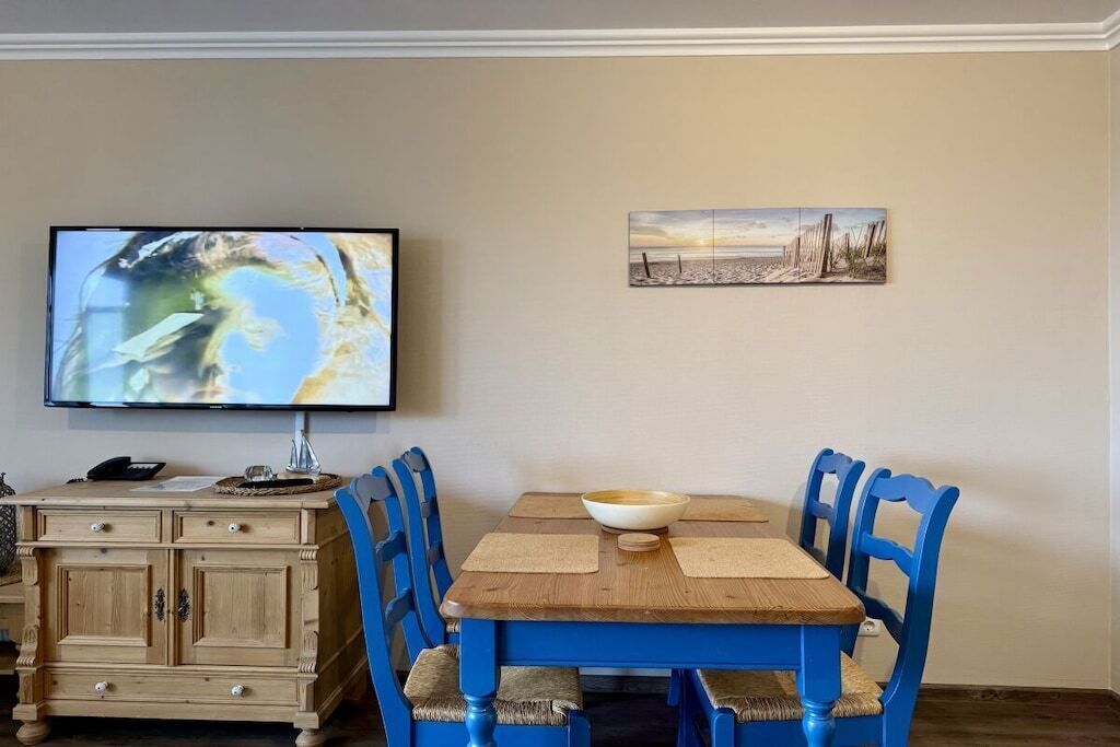 Un área de comedor con una mesa de madera y sillas azules, un televisor montado en la pared y un mueble bajo con cajones.