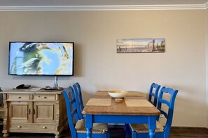 A dining area with a wooden table and blue chairs, a television mounted on the wall, and a small cabinet with drawers. - Holiday apartment with 1 bedroom (Travemünde)